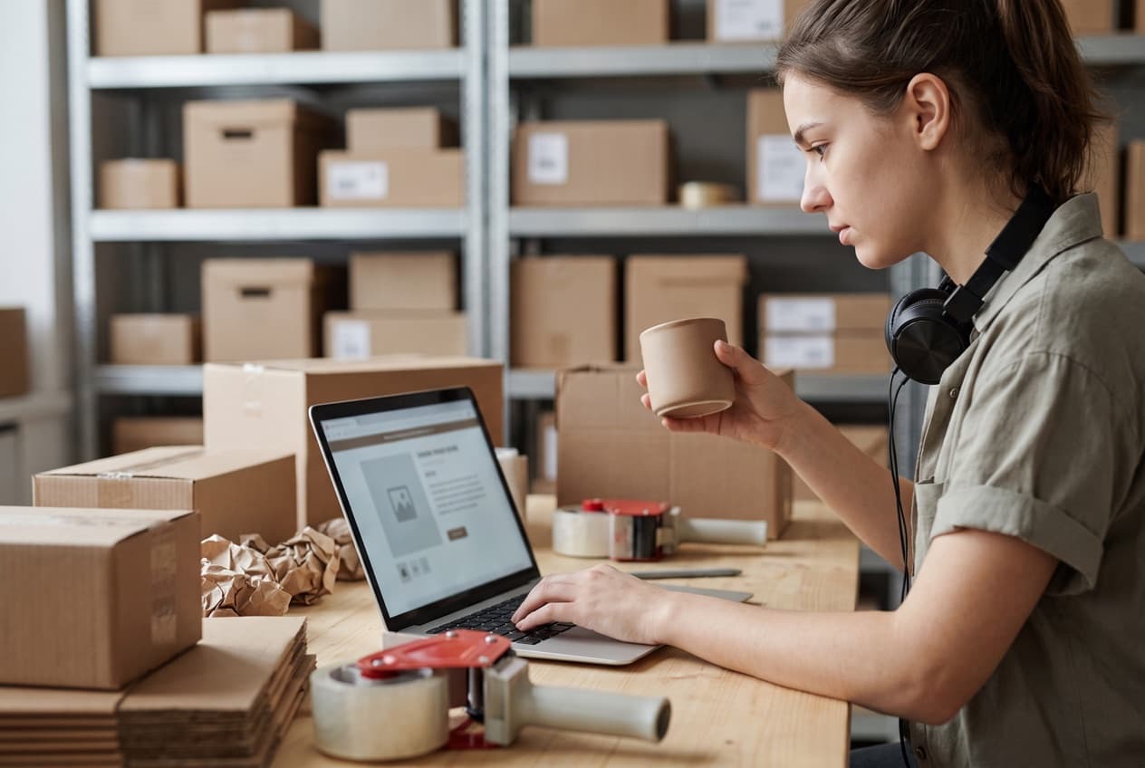 Responsable de catalogue rédigeant elle-même la description d'un produit sur son ordinateur, au milieu des cartons de préparation