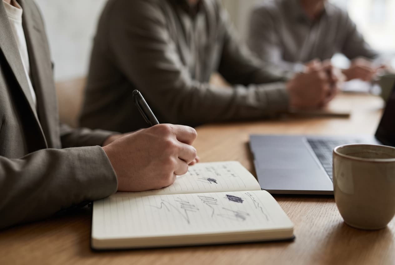 Main qui note quelques lignes au stylo sur un carnet ouvert pendant une réunion de cadrage, le geste par lequel une stratégie social ads pose son hypothèse et son critère d'échec avant le lancement